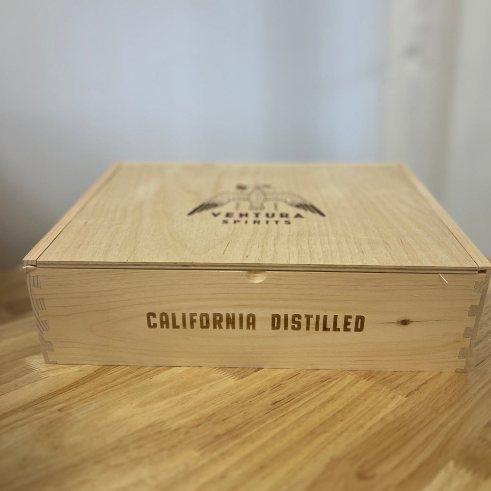 A beige wooden gift box with 'California Distilled' text and a laser engraved logo of Ventura Spirits on it, placed on a wooden surface.