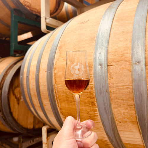 A glass with Strawberry Thief branding held up tp a cast from which the brandy is aged.