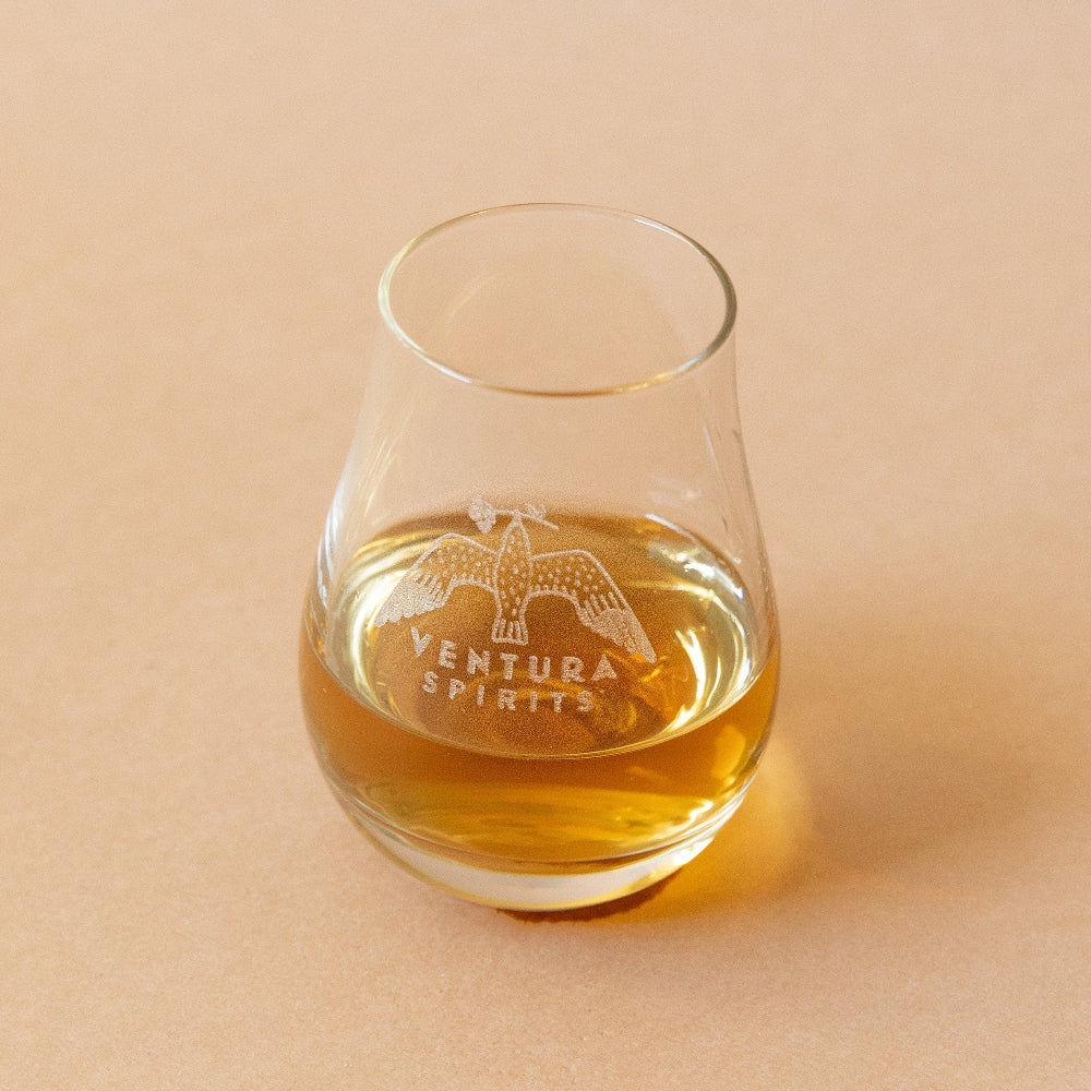 A clear glass with a golden liquid and the Ventura Spirits logo etched on the side, placed on a beige background.